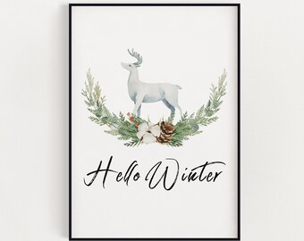 May include: A framed watercolor print featuring a white deer standing above a wreath of greenery, cotton, and a pinecone. The words "Hello Winter" are written in elegant script below the wreath. The artwork is set against a white background.