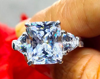 May include: A radiant, silver ring featuring a large, square-cut, clear gemstone as the centerpiece, flanked by two smaller, triangular stones. The ring's setting is silver, and the gemstones sparkle brightly against a blurred red background.
