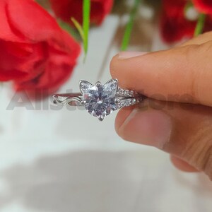 May include: A silver ring with a cat-shaped design. The ring features a large, clear gemstone at the centre, surrounded by smaller stones. The band has a delicate, wavy pattern. The ring is held by a hand, with red flowers in the background.