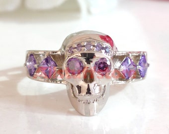 May include: A silver ring with a skull design. The skull has purple and pink gemstones for eyes and a row of smaller purple gemstones around the top of the skull.