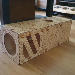 Op de afbeelding: Een houten kattentunnel met een fantasierijk ontwerp met verschillende illustraties. De tunnel heeft twee openingen en een uitsparing aan de bovenkant. De tunnel is gelabeld met "Aslan & Ahsoka".