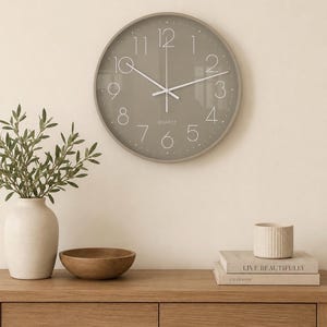 May include: A round wall clock with a taupe face, white numerals, and white hands. The word QUARTZ is printed in white below the centre. The clock features a thin frame and a glass cover, displaying a clear numerical layout from one to twelve.