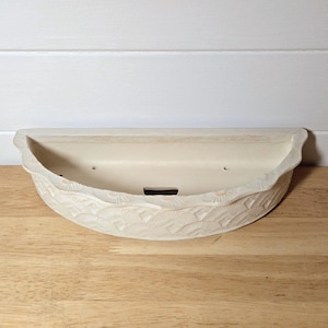 May include: Off-white ceramic wall planter with a curved design. The planter has a textured, wave-like pattern along the bottom edge and a smooth interior. It features two small drainage holes and a rectangular opening at the base.