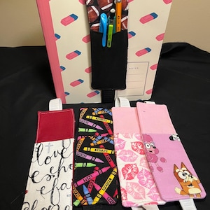 May include: Four rectangular fabric pen holders with elastic bands designed to attach to notebooks. One features a football pattern, one has white fabric with black cursive text, one displays colorful crayons on black, and one shows pink lips and cartoon dogs.