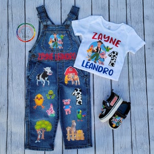 Peut inclure: Un ensemble de salopette en jean avec un t-shirt blanc assorti et des baskets noires. La salopette et les chaussures pr&eacute;sentent des graphiques de dessins anim&eacute;s d'animaux de la ferme et le nom "Zayne Leandro". Le t-shirt porte le nom "Zayne" et le chiffre "1".