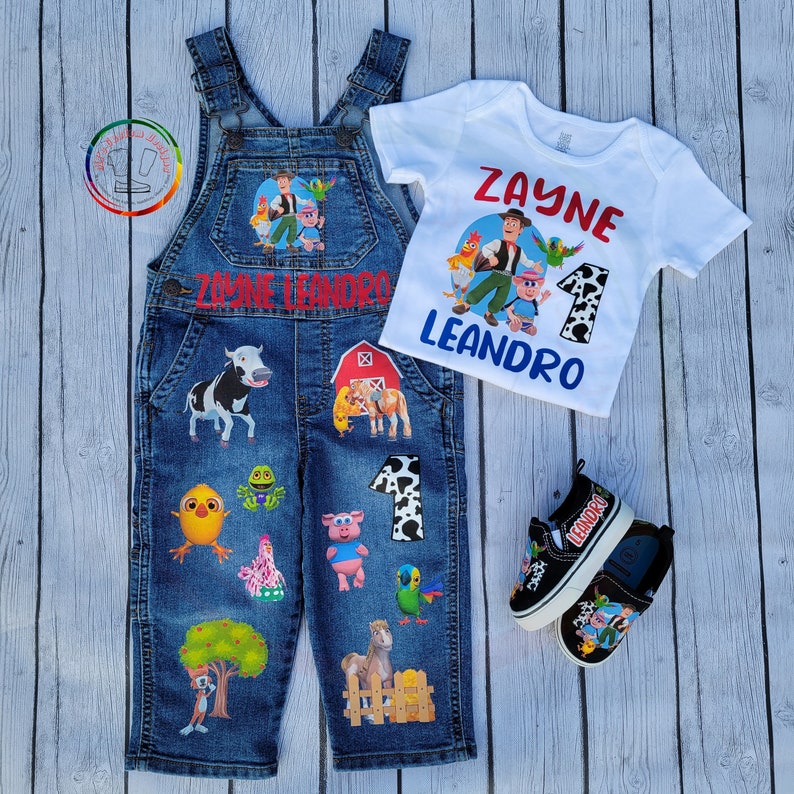 Peut inclure: Un ensemble de salopette en jean avec un t-shirt blanc assorti et des baskets noires. La salopette et les chaussures pr&eacute;sentent des graphiques de dessins anim&eacute;s d'animaux de la ferme et le nom "Zayne Leandro". Le t-shirt porte le nom "Zayne" et le chiffre "1".