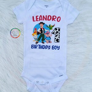 Peut inclure: Body b&eacute;b&eacute; blanc avec un graphisme color&eacute; repr&eacute;sentant un cow-boy de dessin anim&eacute; et d'autres personnages. Le texte "LEANDRO" en rouge et "BIRTHDAY BOY" en bleu est imprim&eacute; sur le devant, avec le chiffre "1".