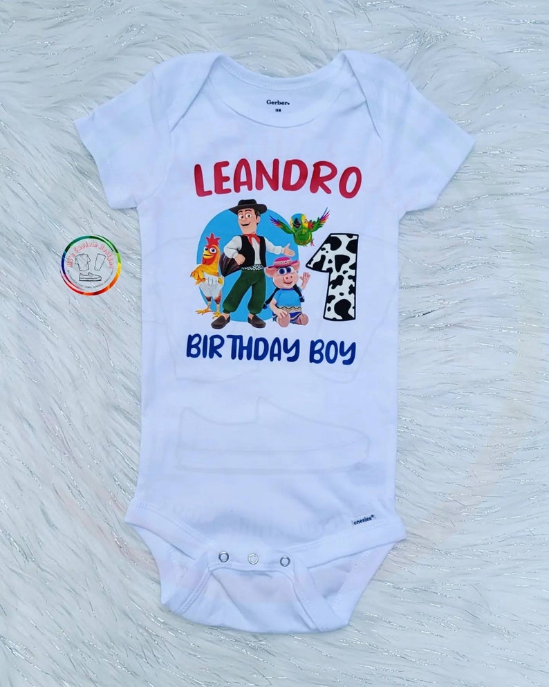 Peut inclure: Body b&eacute;b&eacute; blanc avec un graphisme color&eacute; repr&eacute;sentant un cow-boy de dessin anim&eacute; et d'autres personnages. Le texte "LEANDRO" en rouge et "BIRTHDAY BOY" en bleu est imprim&eacute; sur le devant, avec le chiffre "1".