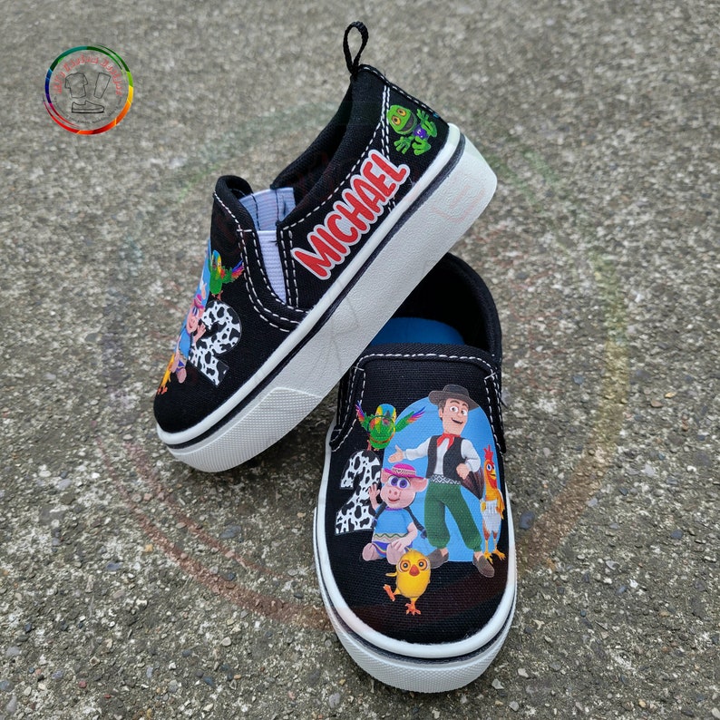 Peut inclure: Chaussures &agrave; enfiler noires avec des personnages de dessins anim&eacute;s color&eacute;s et le nom "MICHAEL" en rouge. Les chaussures ont des semelles blanches et une tige noire, avec une tirette au talon. Le design comprend divers personnages et le chiffre 2.