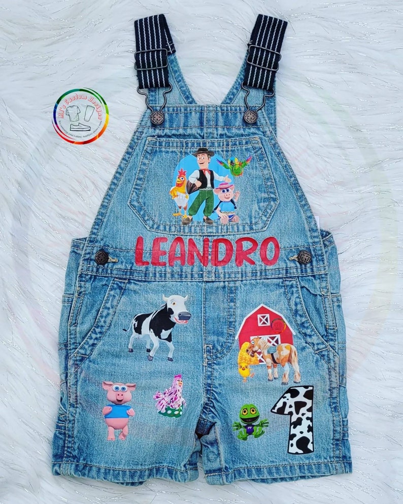 K&ouml;nnte beinhalten: Blaue Jeans-Latzhose mit Applikationen von Cartoon-Bauernhoftieren und -figuren. Der Name "LEANDRO" steht in roten Buchstaben auf der Vorderseite. Die Latzhose hat verstellbare Tr&auml;ger und einen Aufn&auml;her mit der Zahl "1".