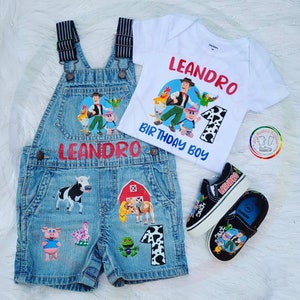 K&ouml;nnte beinhalten: Ein Denim-Latzhosen-Shorts-Set mit passendem wei&szlig;em T-Shirt und schwarzen Turnschuhen. Das Set zeigt Cartoon-Bauernhoftier- und Charaktergrafiken, den Namen "LEANDRO" und den Text "BIRTHDAY BOY". Die Turnschuhe tragen den Namen "LEANDRO".