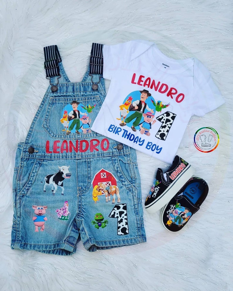 K&ouml;nnte beinhalten: Ein Denim-Latzhosen-Shorts-Set mit passendem wei&szlig;em T-Shirt und schwarzen Turnschuhen. Das Set zeigt Cartoon-Bauernhoftier- und Charaktergrafiken, den Namen "LEANDRO" und den Text "BIRTHDAY BOY". Die Turnschuhe tragen den Namen "LEANDRO".