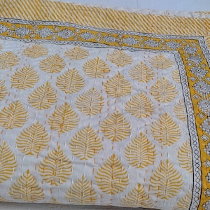 May include: A white and yellow quilt with a leaf pattern. The quilt has a yellow border with a striped design and a decorative border with geometric shapes. The quilt is made of a soft material and is likely handmade.