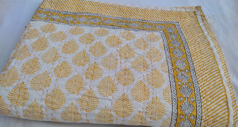 May include: A white and yellow quilt with a leaf pattern. The quilt has a yellow border with a striped design and a decorative border with geometric shapes. The quilt is made of a soft material and is likely handmade.