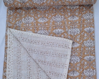 May include: A rolled-up, tan-colored quilt with a floral pattern in white. The quilt is partially unfolded, revealing a cream-colored interior with stitched lines. The floral design features stylized flowers and leaves.