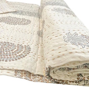 May include: A cream-colored cotton quilt featuring a repeating block-printed pattern of dark gray and brown paisley-style floral motifs. The textile is finished with visible horizontal running stitches in a light brown thread throughout the surface.