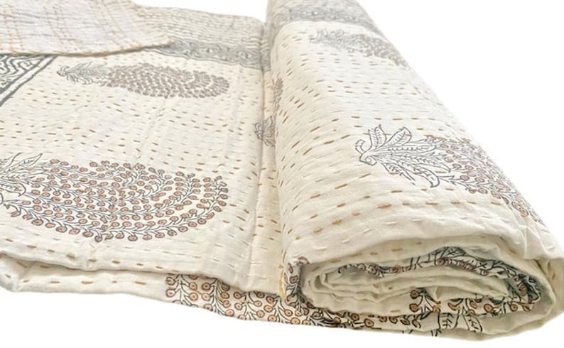 May include: A cream-colored cotton quilt featuring a repeating block-printed pattern of dark gray and brown paisley-style floral motifs. The textile is finished with visible horizontal running stitches in a light brown thread throughout the surface.