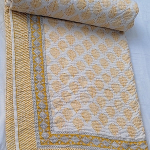 May include: A rolled-up, off-white quilt with a repeating pattern of yellow leaf shapes. The quilt has a yellow and white striped border and a decorative border with geometric designs. The quilt is folded and ready for use.