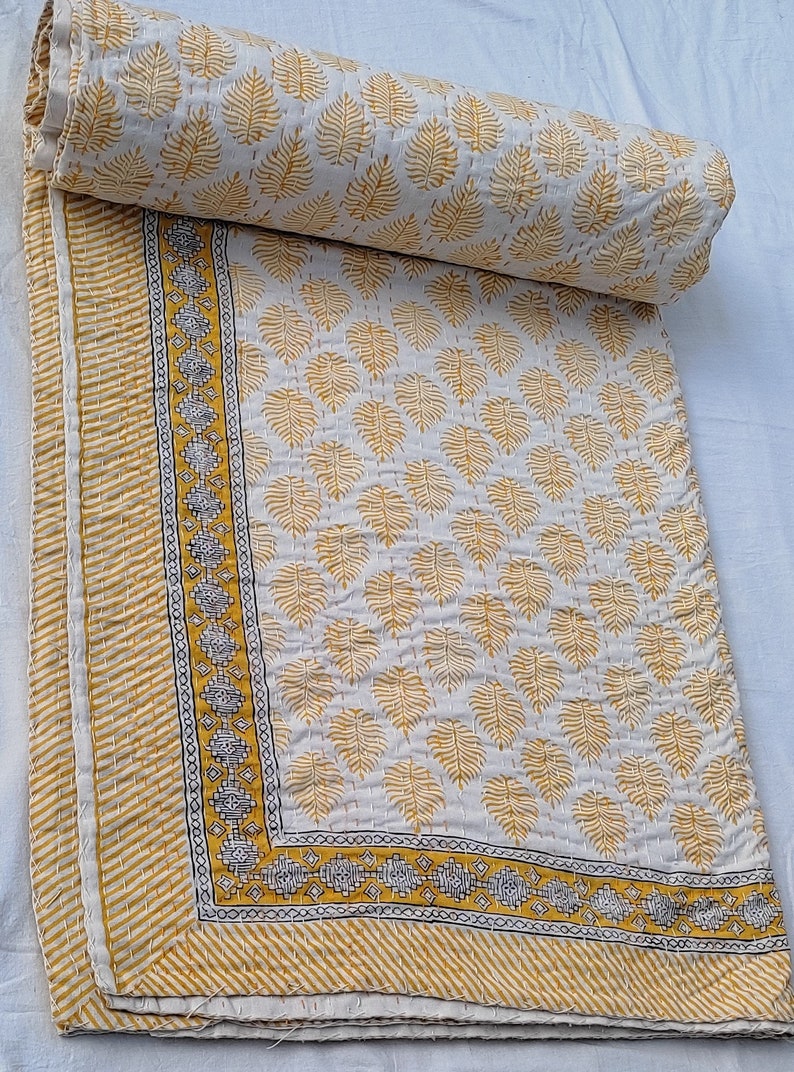 May include: A rolled-up, off-white quilt with a repeating pattern of yellow leaf shapes. The quilt has a yellow and white striped border and a decorative border with geometric designs. The quilt is folded and ready for use.