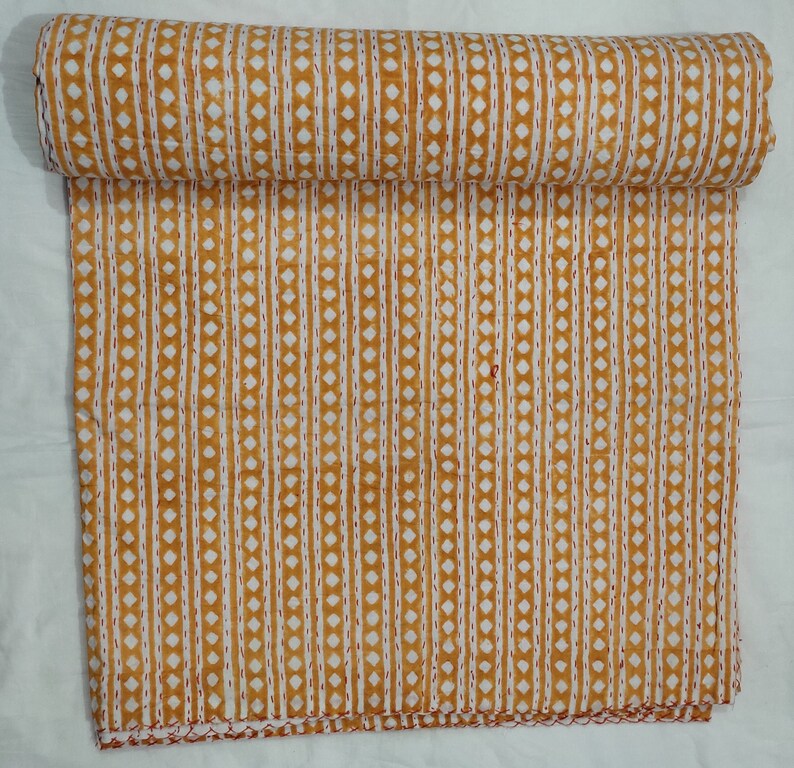 May include: A yellow and white kantha quilt with a repeating pattern of small diamonds and vertical lines. The quilt is folded in half and is lying on a white surface.
