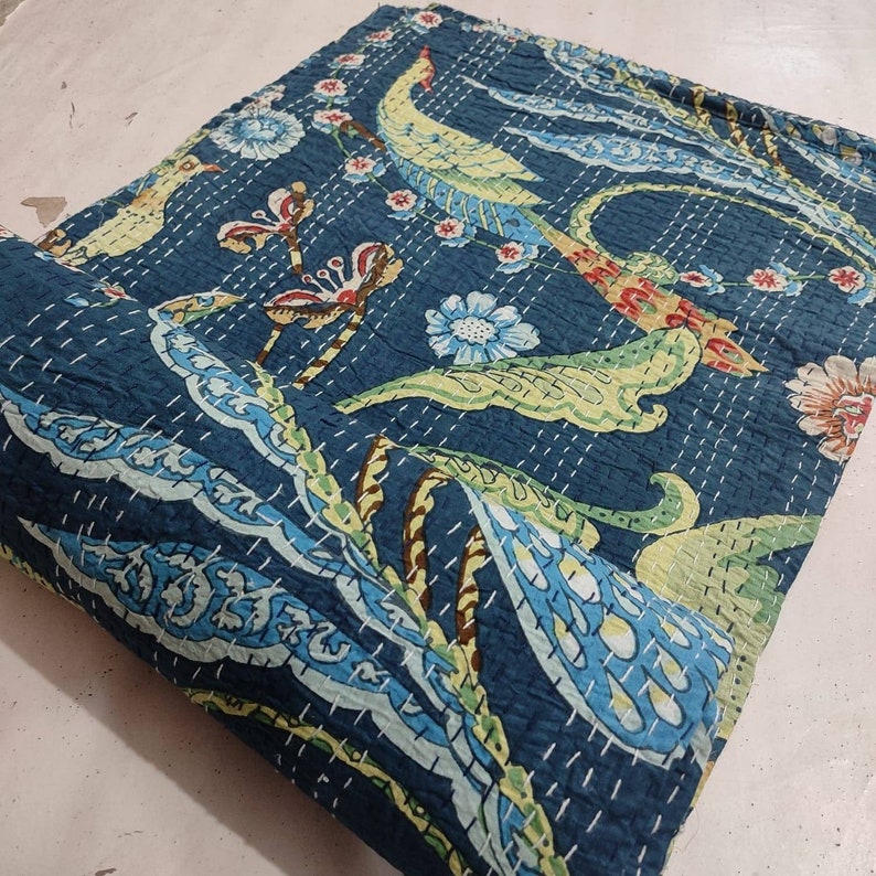 May include: A dark blue Kantha quilt with a repeating pattern of birds, flowers, and foliage in shades of yellow, green, red, and blue. The quilt is hand-stitched with white thread, creating a textured surface. The edges are neatly finished.