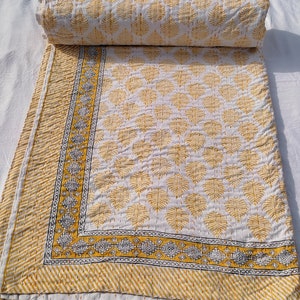 May include: A white and yellow quilt with a leaf pattern. The quilt is folded and rolled up, with the leaf pattern visible on the unfolded portion. The border features a geometric design and diagonal stripes. The quilt is made of a soft, quilted material.