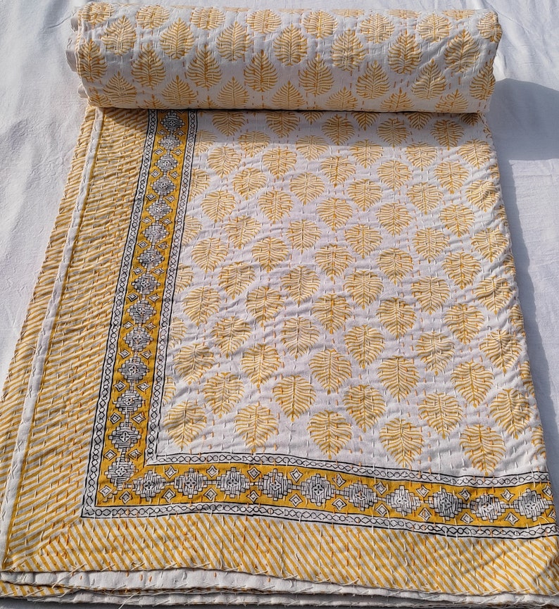 May include: A white and yellow quilt with a leaf pattern. The quilt is folded and rolled up, with the leaf pattern visible on the unfolded portion. The border features a geometric design and diagonal stripes. The quilt is made of a soft, quilted material.