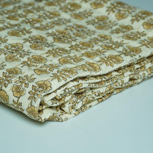 Op de afbeelding: Gevouwen crèmekleurige quilt met een herhalend bloemenpatroon in goud- en bruintinten. De quilt van zachte stof heeft een gewatteerd ontwerp. Het bloemenpatroon omvat bloemen en bladeren.