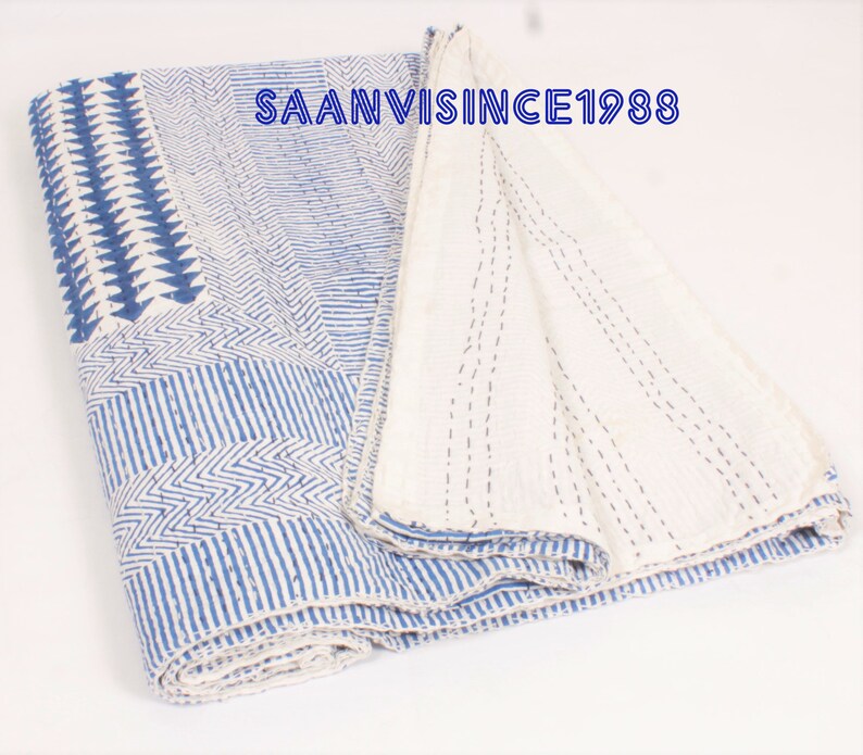 May include: A folded cotton quilt featuring a blue and white geometric pattern with visible running stitch details. The fabric displays various linear, chevron, and triangular motifs. The text SAANVISINCE1988 is printed in blue on the quilt surface.