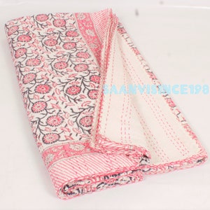 Op de afbeelding: Een gevouwen quilt met een bloemenpatroon, een crèmekleurige achtergrond en roze en zwarte bloemmotieven. De quilt heeft een roze rand met een streepjespatroon en is met roze draad met de hand gestikt. De tekst "SAANVISINCE1988" is zichtbaar.