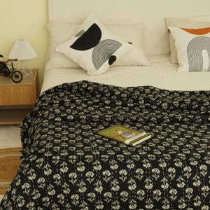 May include: A black quilt featuring a repeating white floral pattern covers a bed with white pillows, including two decorative pillows with black and orange geometric designs, and a small book resting on the surface of the quilt.
