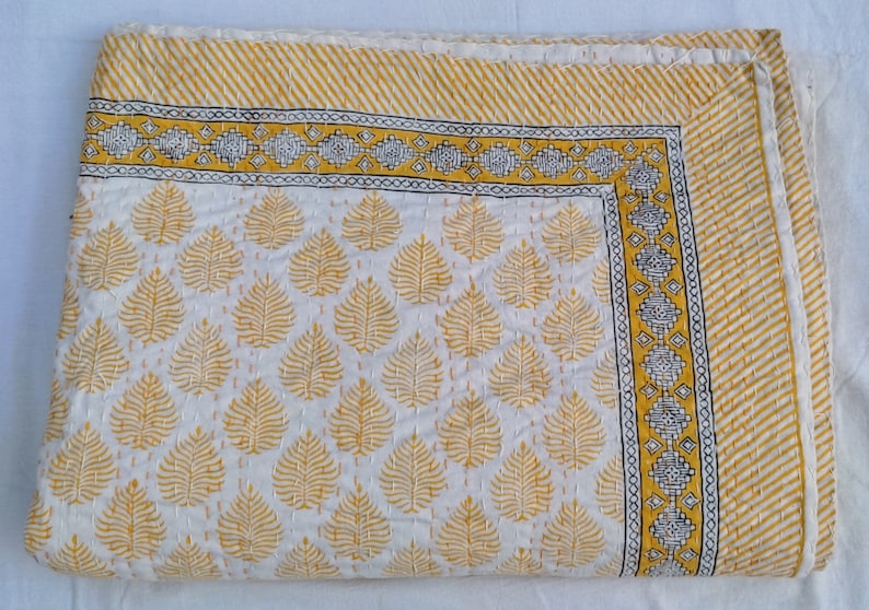 May include: A white and yellow quilt with a leaf pattern. The quilt features a repeating pattern of yellow leaves on a white background. The border has a yellow and white striped design with a decorative yellow and black border.