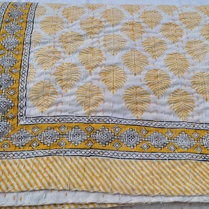 May include: A folded white and yellow quilt with a leaf pattern. The quilt features a border with geometric designs and a striped edge. The leaf pattern is in yellow, and the border has black and white accents.