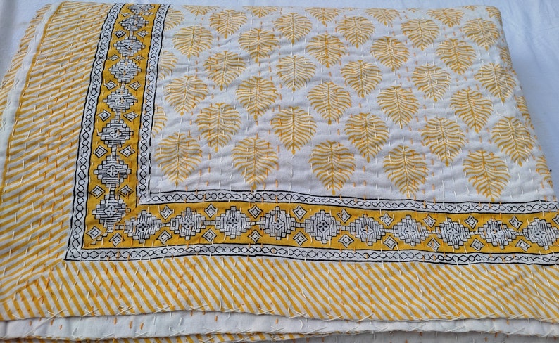 May include: A folded white and yellow quilt with a leaf pattern. The quilt features a border with geometric designs and a striped edge. The leaf pattern is in yellow, and the border has black and white accents.