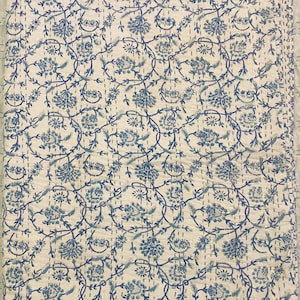 May include: A folded, white quilt with a repeating pattern of blue floral vines and flowers. The quilt is stitched with a contrasting thread, and the edges are scalloped. The overall design is a classic, vintage style.