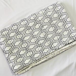 May include: A folded white quilt featuring a repeating black geometric pattern of interlocking curved shapes. The fabric displays visible linear stitching throughout, creating a textured appearance across the entire surface of the textile.