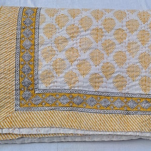 May include: Folded white and yellow quilt with a leaf pattern. The quilt features a yellow border with a geometric design and a striped edge. The leaf pattern is in yellow, and the stitching is visible.