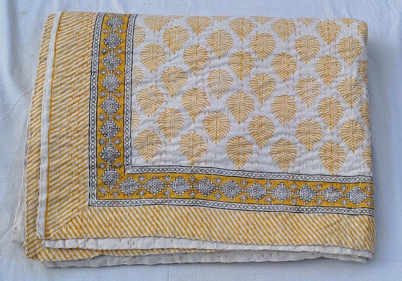 May include: Folded white and yellow quilt with a leaf pattern. The quilt features a yellow border with a geometric design and a striped edge. The leaf pattern is in yellow, and the stitching is visible.