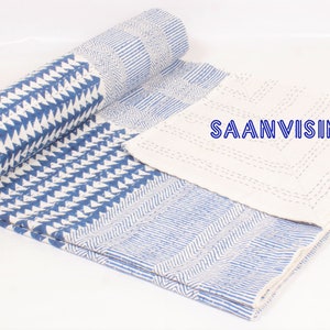 May include: A partially rolled quilt featuring a blue and white geometric pattern with triangular and linear motifs. The fabric displays visible white stitching throughout, and the text SAANVISINCE1988 is printed in blue on the white section.
