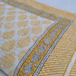 May include: A folded white and yellow quilt with a leaf pattern. The quilt has a yellow and white striped border and a decorative border with black and yellow geometric designs. The quilt is hand-stitched.