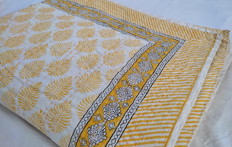 May include: A folded white and yellow quilt with a leaf pattern. The quilt has a yellow and white striped border and a decorative border with black and yellow geometric designs. The quilt is hand-stitched.