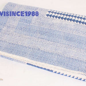 May include: A folded cotton quilt featuring a white base with blue block-printed patterns including chevron stripes, parallel lines, and triangles. The text SAANVISINCE1988 is printed in blue on the upper left corner of the fabric surface.
