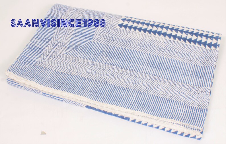 May include: A folded cotton quilt featuring a white base with blue block-printed patterns including chevron stripes, parallel lines, and triangles. The text SAANVISINCE1988 is printed in blue on the upper left corner of the fabric surface.