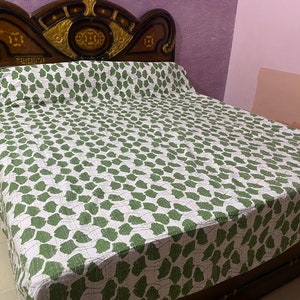 May include: A bedspread featuring a repeating pattern of green leaf-like shapes on a white background. The bedspread is draped over a bed with a dark brown wooden headboard that has gold-colored decorative accents and text in a non-Latin script.