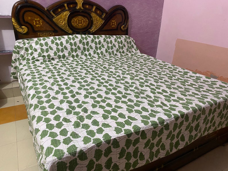 May include: A bedspread featuring a repeating pattern of green leaf-like shapes on a white background. The bedspread is draped over a bed with a dark brown wooden headboard that has gold-colored decorative accents and text in a non-Latin script.