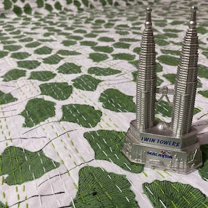 May include: A silver-colored metal souvenir figurine of the Petronas Twin Towers. The base features the text "TWIN TOWERS" and "MALAYSIA" in blue lettering, with embossed architectural details around the perimeter of the stand.