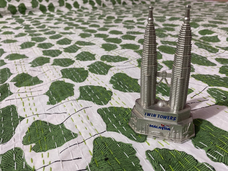 May include: A silver-colored metal souvenir figurine of the Petronas Twin Towers. The base features the text "TWIN TOWERS" and "MALAYSIA" in blue lettering, with embossed architectural details around the perimeter of the stand.