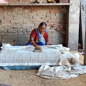 May include: A person uses a wooden block to stamp a dark brown geometric pattern onto a white fabric sheet. The fabric features a repeating circular and triangular design, and the person wears a red shirt with a blue and white patterned headscarf.