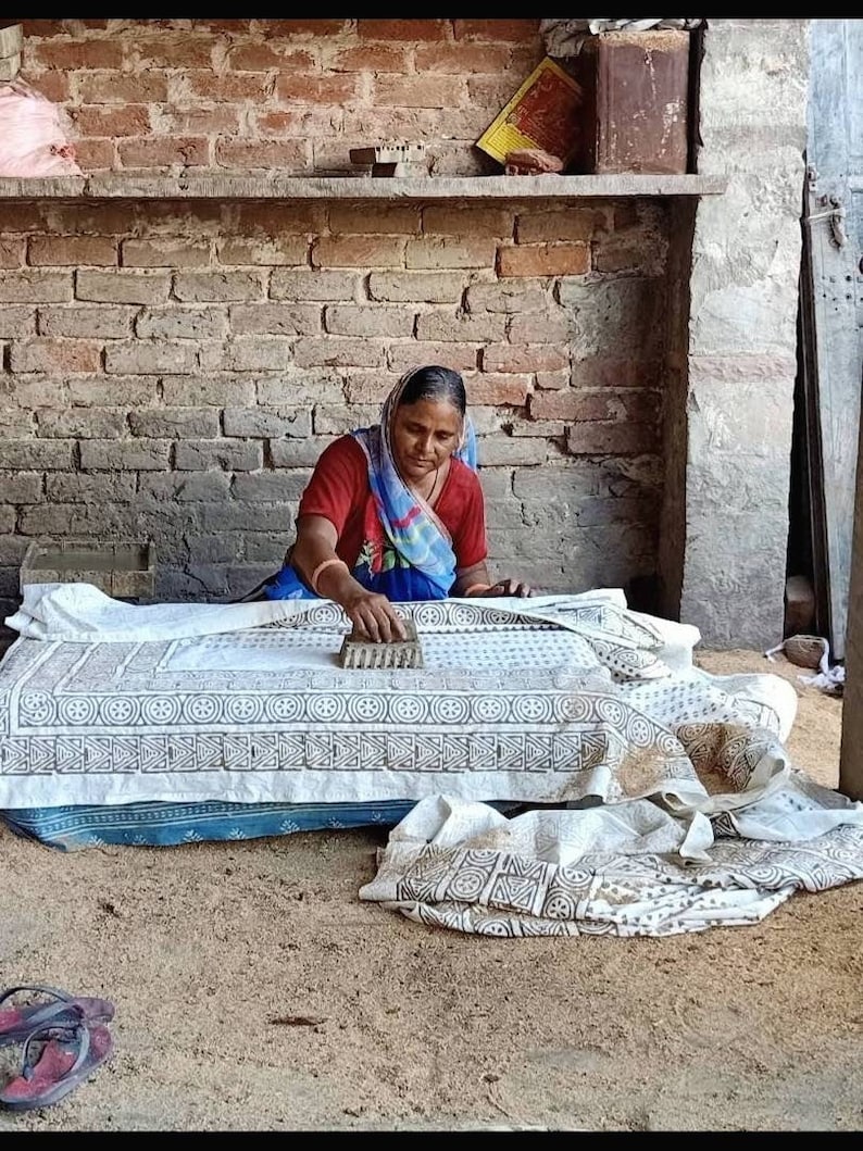 May include: A person uses a wooden block to stamp a dark brown geometric pattern onto a white fabric sheet. The fabric features a repeating circular and triangular design, and the person wears a red shirt with a blue and white patterned headscarf.