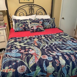 May include: A bed with a navy blue quilt and matching pillow shams featuring a colorful bird and floral design. The bed also has a red bedspread and white pillows. The bed frame is black.
